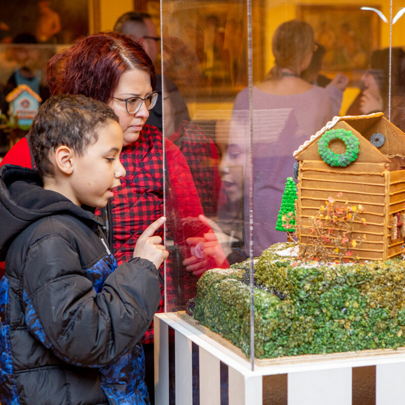 A child and an adult are viewing one of many spectaculare gingerbread houses featured at The Gingerbread Invitational.